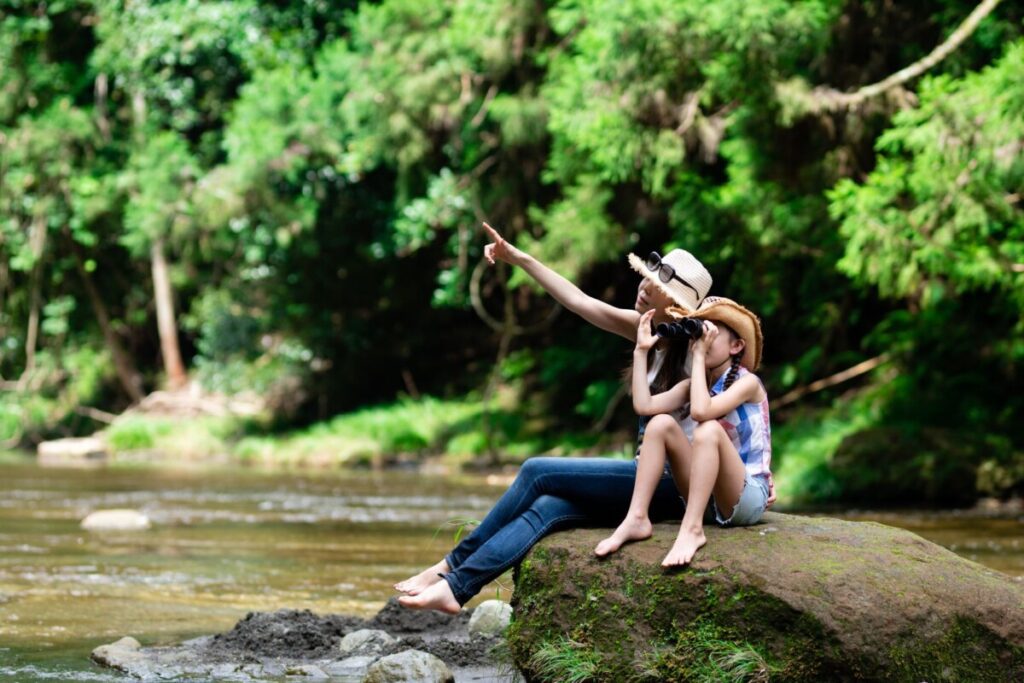 A mother and daughter use binoculars sitting near a forest creek.