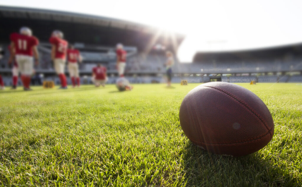 A football sits on the grass with football players in the background